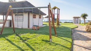 A playground with swings and a swing set, a thatched-roof building, and a stone pathway leading to a gate. - Mi Capricho Beach Front sea View Pool (Mijas)