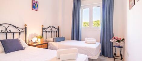 A bedroom with two beds, a nightstand with a lamp, a window with curtains, and a small table with a vase of flowers.