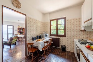 A kitchen with a dining area, wooden chairs, a table with a white tablecloth, a clock on the wall, and a window with a view of greenery. - Lago Azzurro Infinity View (Premeno)