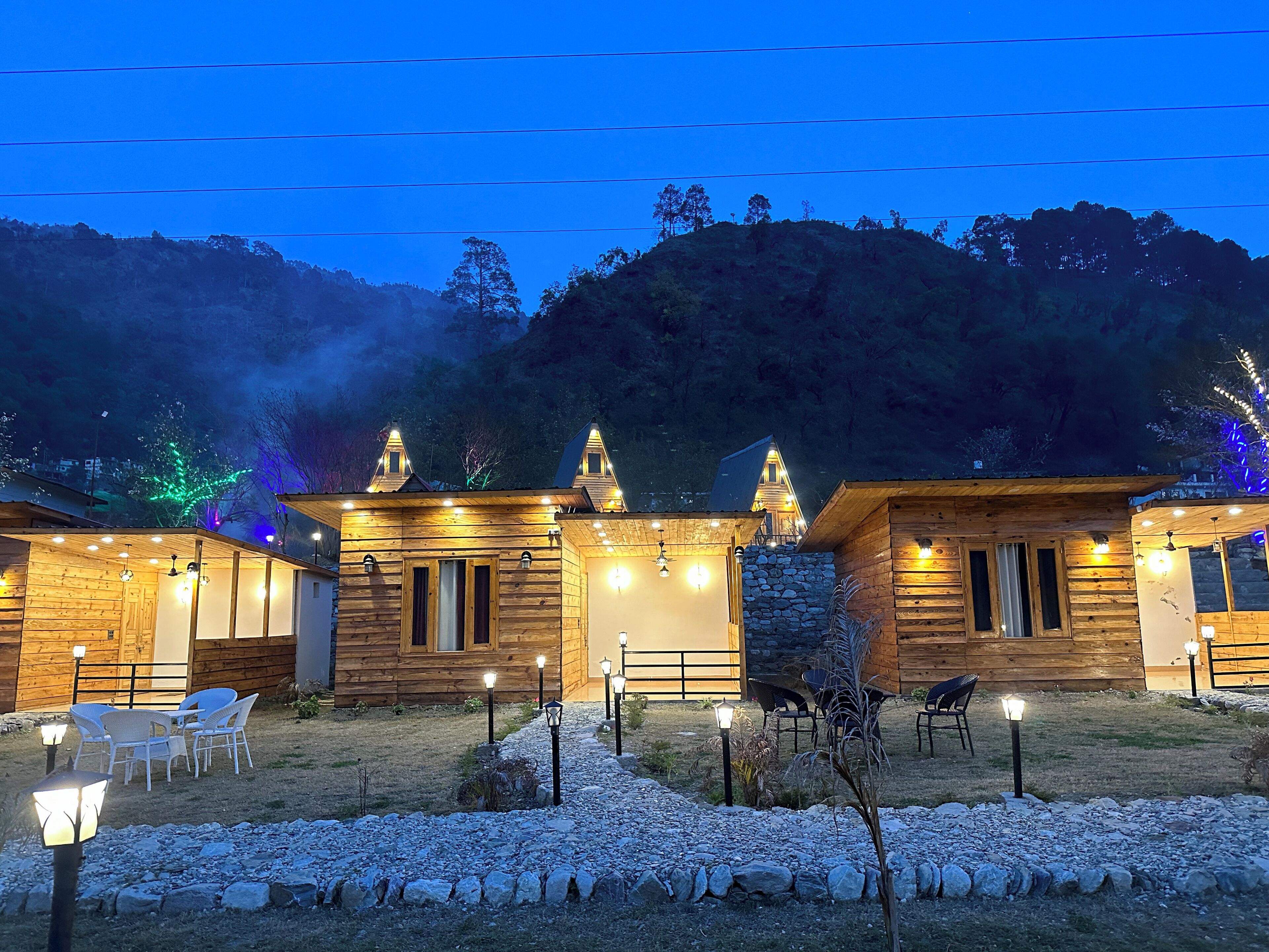 A row of wooden cabins with stone walls, outdoor seating, and a view of a forested hill.