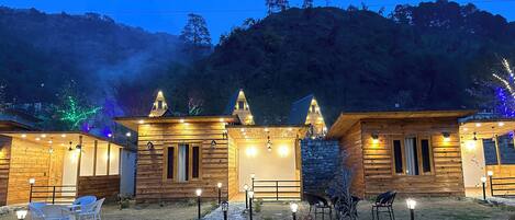 A row of wooden cabins with stone walls, outdoor seating, and a view of a forested hill.