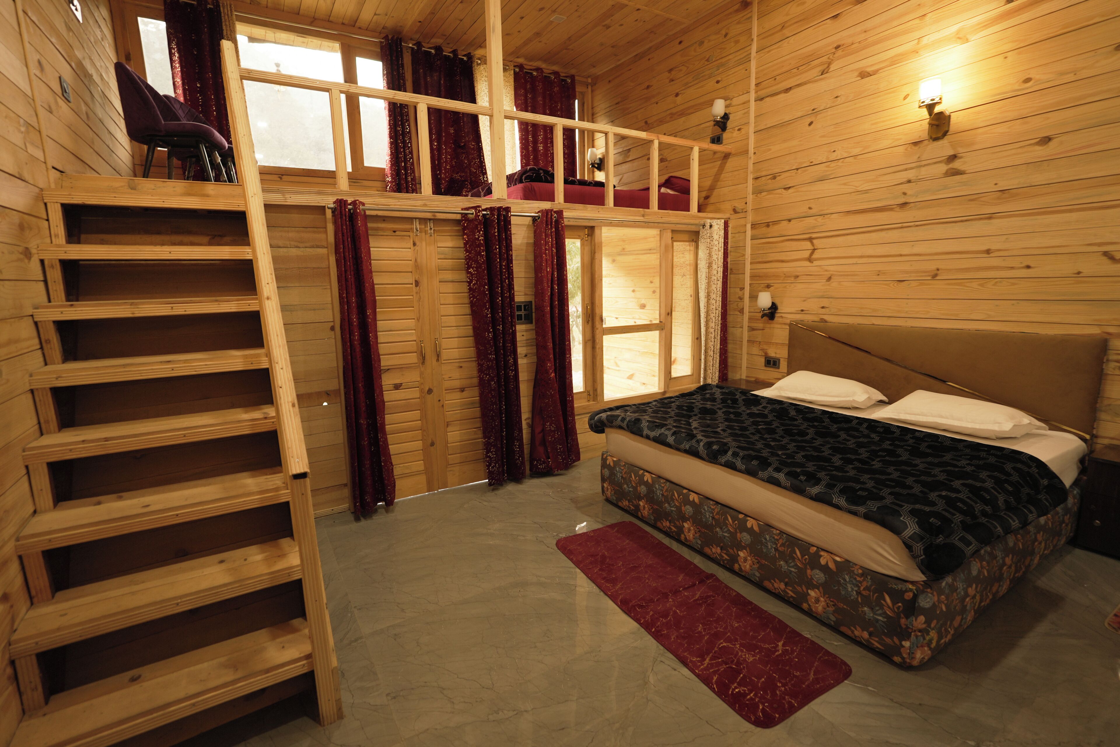 Un dormitorio de cabaña de madera con literas, una escalera, una ventana con cortinas y una mesita de noche con lámpara.