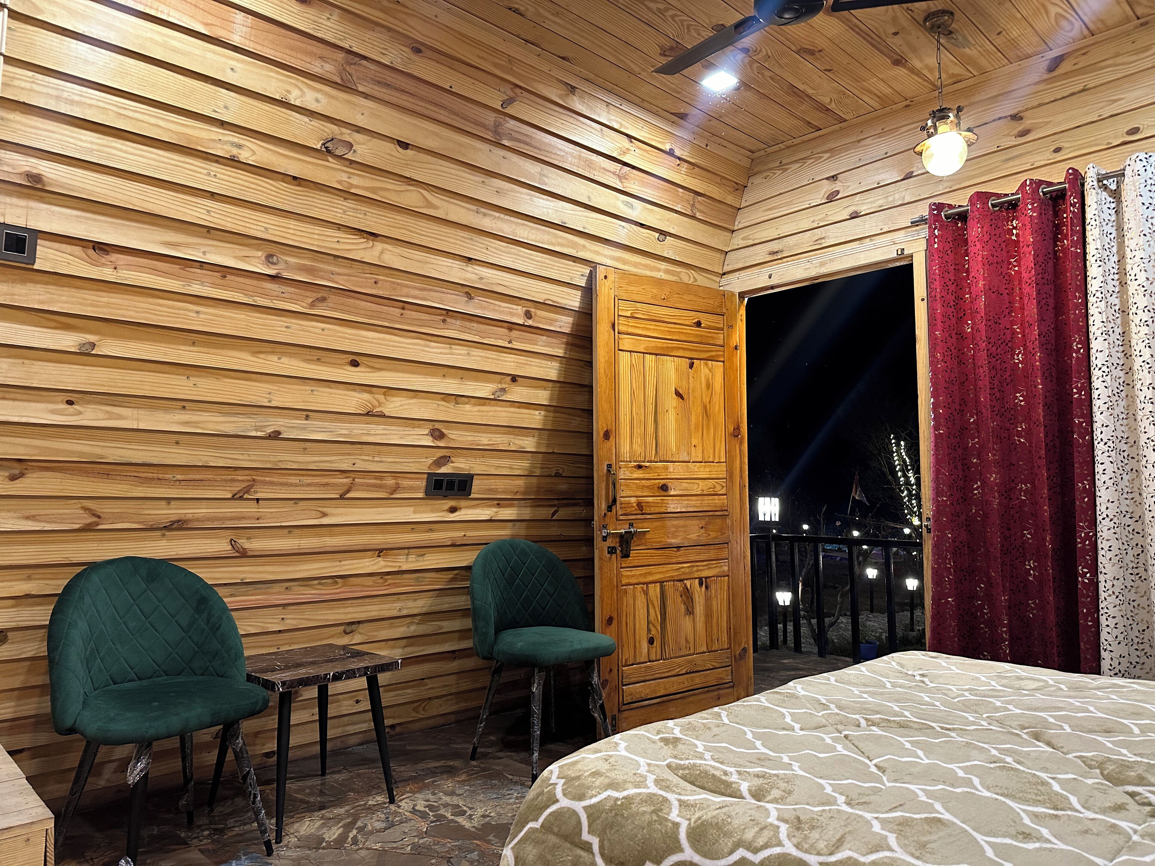 A wooden cabin room with a bed, two chairs, a small table, and a door leading outside.