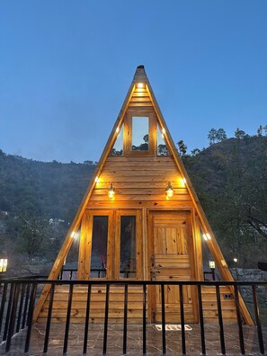A wooden triangular cabin with a central window and two side windows, surrounded by a metal railing. - Cabana Riverside Resort - Kainchi Dham (Kosiyakutoli)