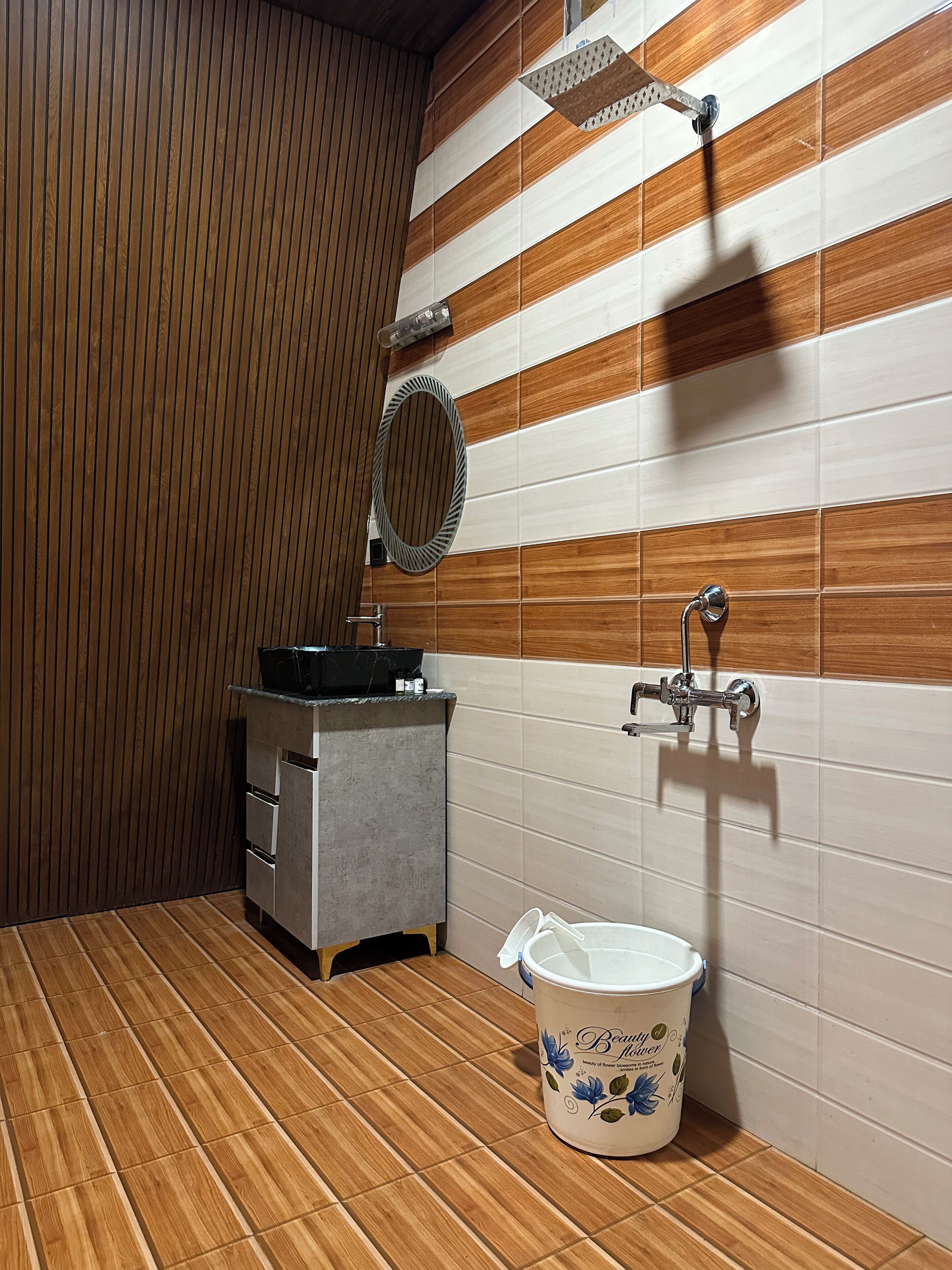 A bathroom with a wooden floor, a sink with a mirror, a shower, and a bucket.