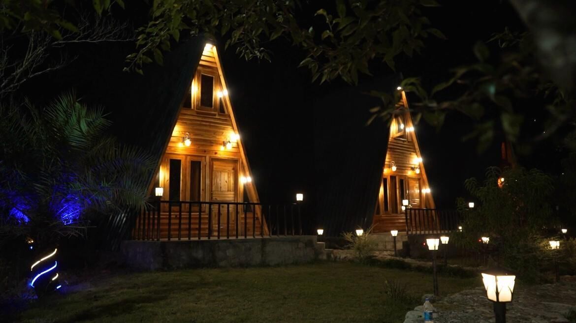 Two wooden cabins with a triangular roof, illuminated by warm lights, surrounded by a landscaped garden with small trees and a pathway.