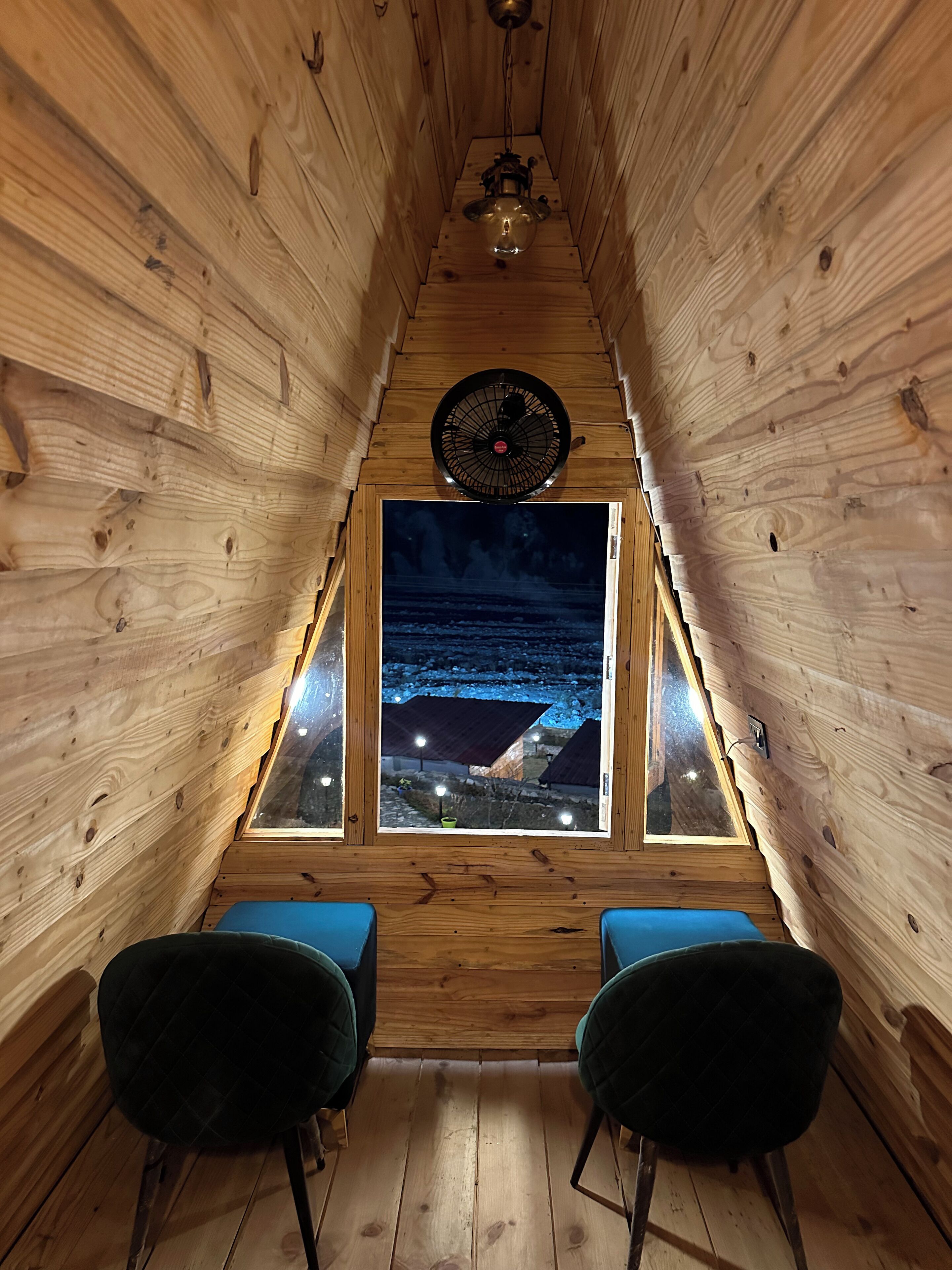 A wooden cabin with a sloped ceiling, a large window, two chairs, and a ceiling fan.
