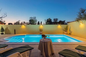 A swimming pool with a table and chairs in the foreground, surrounded by walls with light fixtures. - Villa Monique in Vodnjan (Vodnjan)