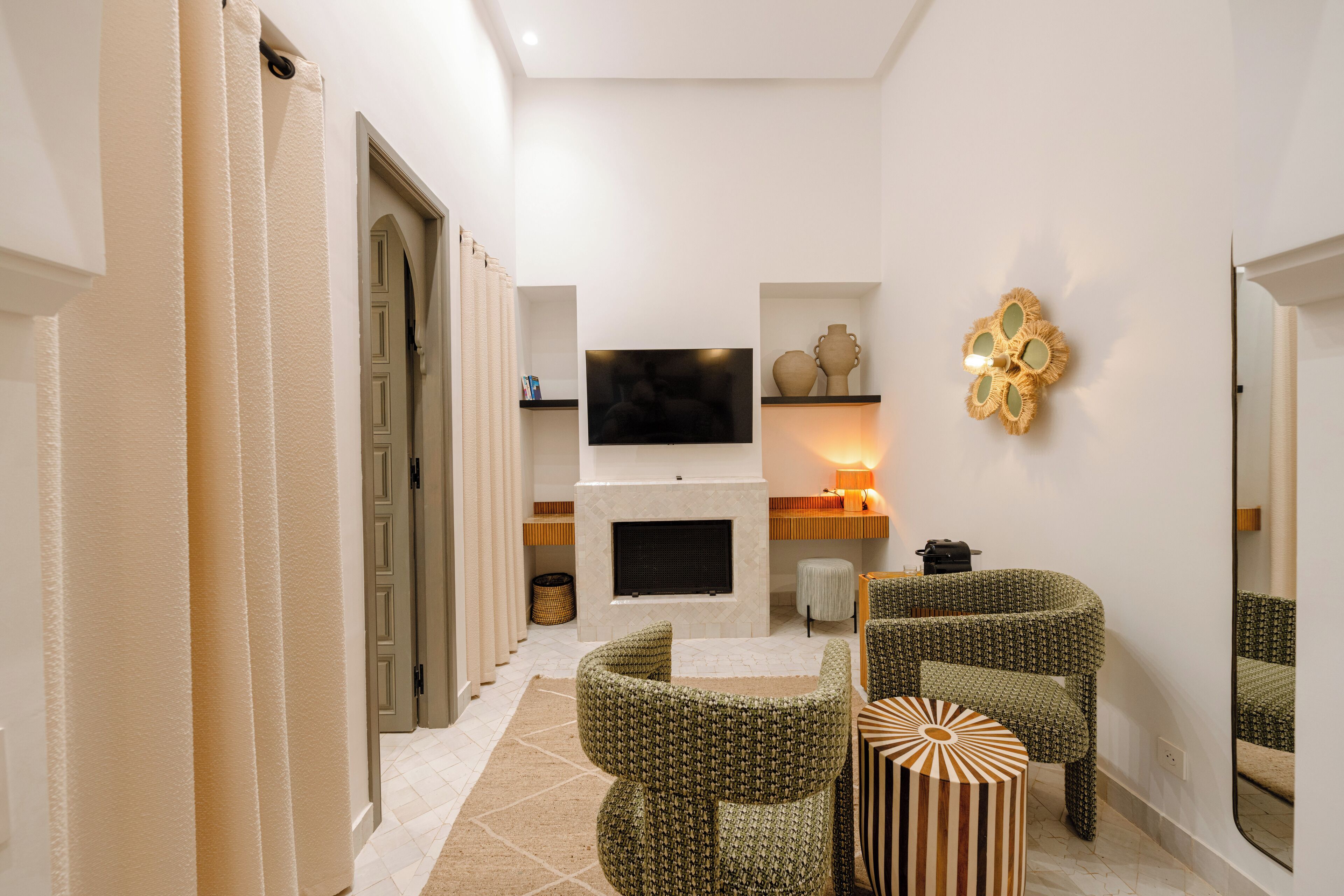 A modern living room with a fireplace, green patterned chairs, a flat-screen TV, and a decorative wall piece.