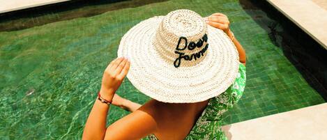 A person in a green and white patterned dress and a straw hat with "Dee Janna" written on it, standing by a pool with clear water.