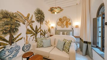 Un dormitorio con un sofá color beige, almohadillas decorativas, una mesa de centro de madera, un mural en la pared y una cama con ropa de cama estampada.