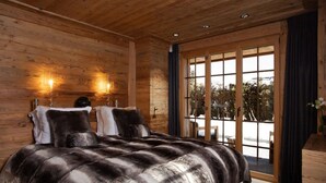 A wooden-paneled bedroom with a large bed, two wall-mounted lights, and a sliding glass door leading to an outdoor area. - Spacious Chalet With Private Pool (Saanen)