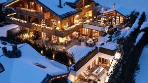 A large, multi-story building with a snow-covered roof and exterior lights. It features multiple balconies and a spacious outdoor patio area. - Spacious Chalet With Private Pool (Saanen)