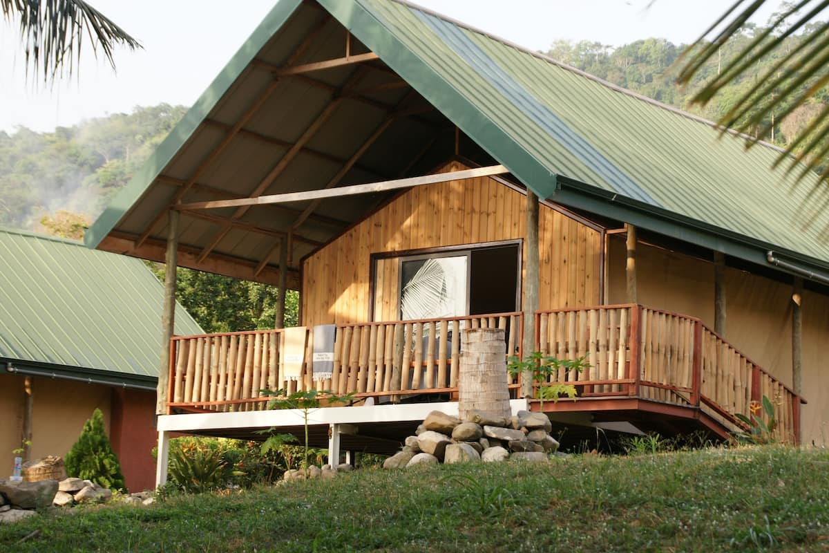 Une cabane en bois dotée d’un balcon, entourée de verdure et avec une chaîne de montagnes en toile de fond.
