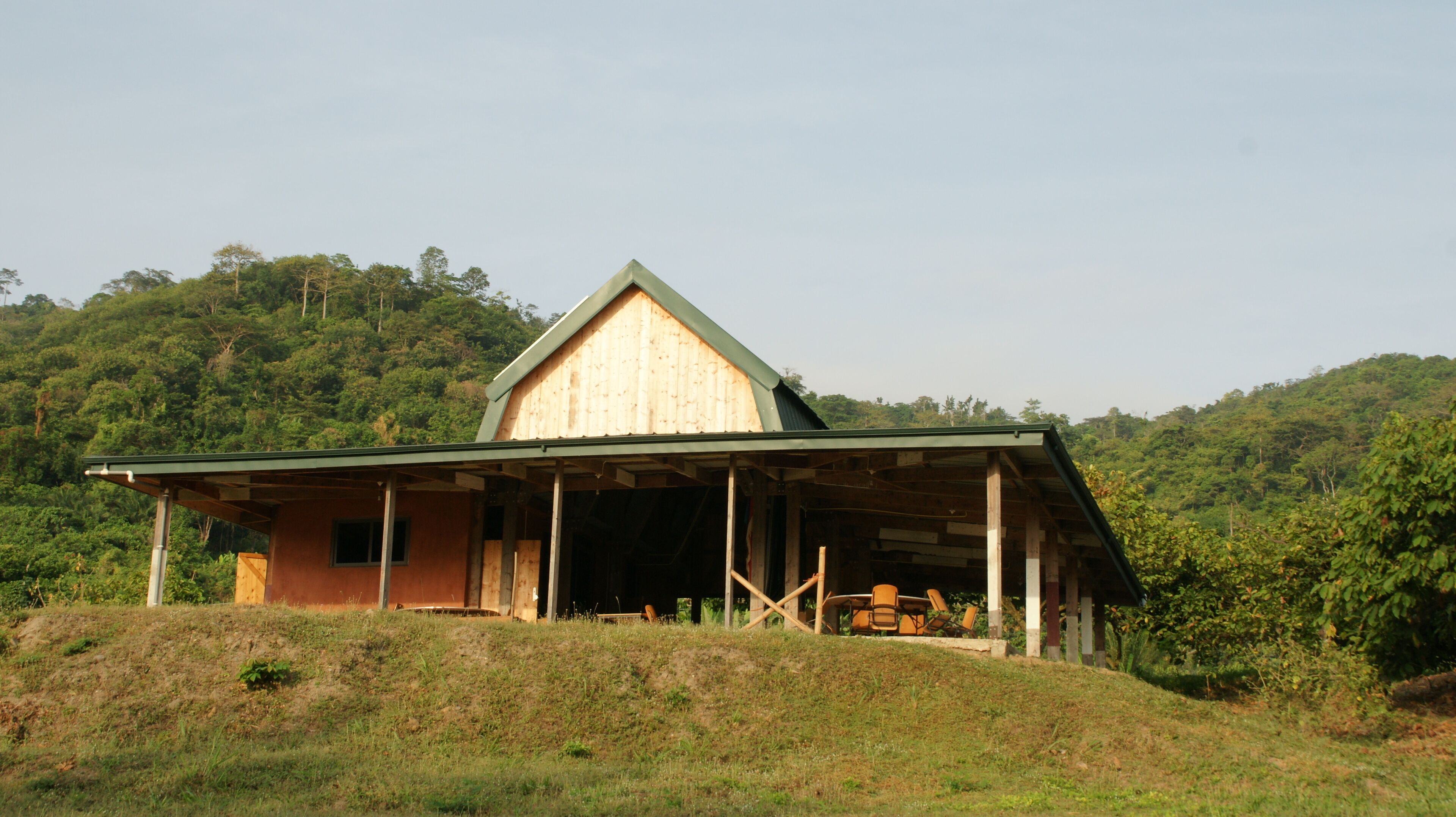 A wooden cabin with a sloped roof, situated on a grassy hill with dense forest in the background.