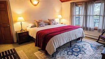 A bedroom with a wooden ceiling, a bed with a red blanket, two bedside lamps, a nightstand, a bench, and a patterned rug.