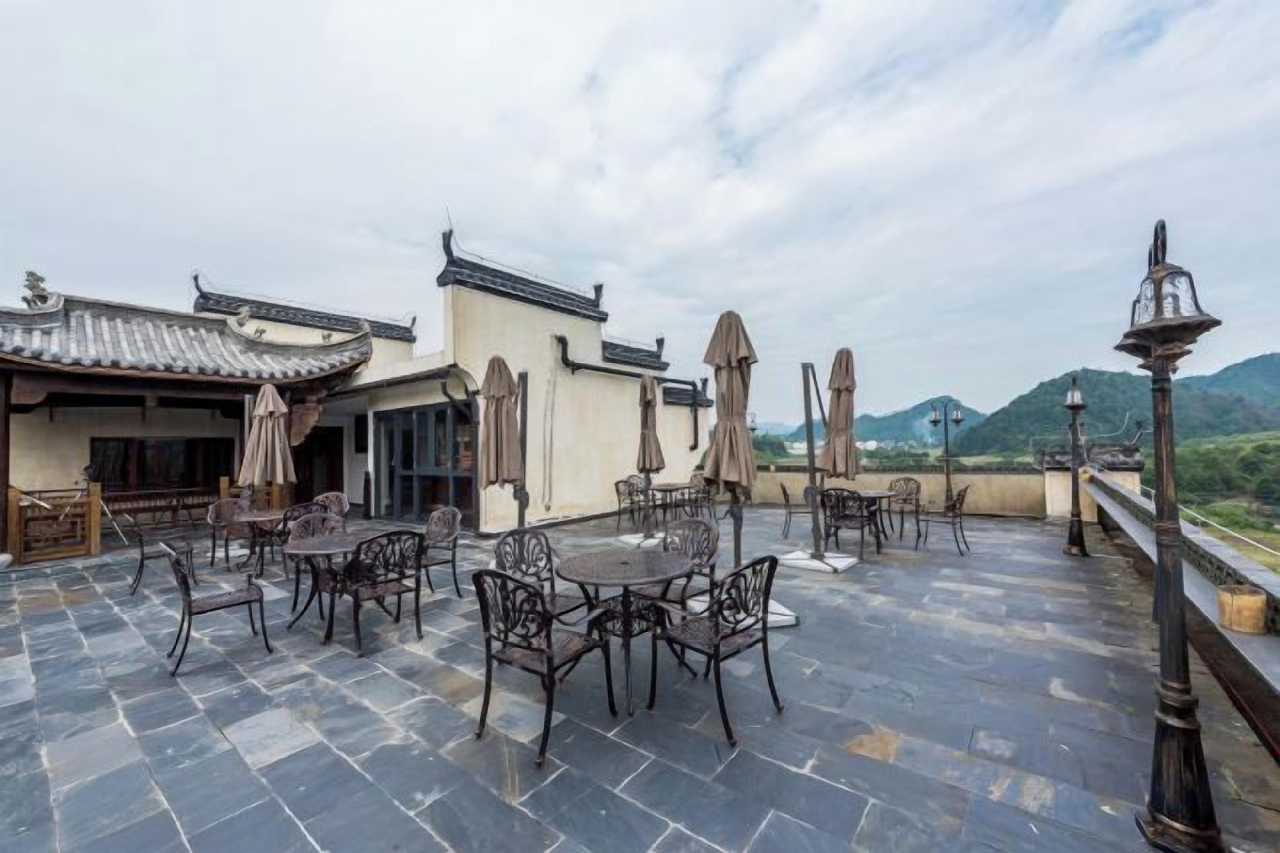 An outdoor patio with tables and chairs, a traditional building with a tiled roof, and a mountainous backdrop.