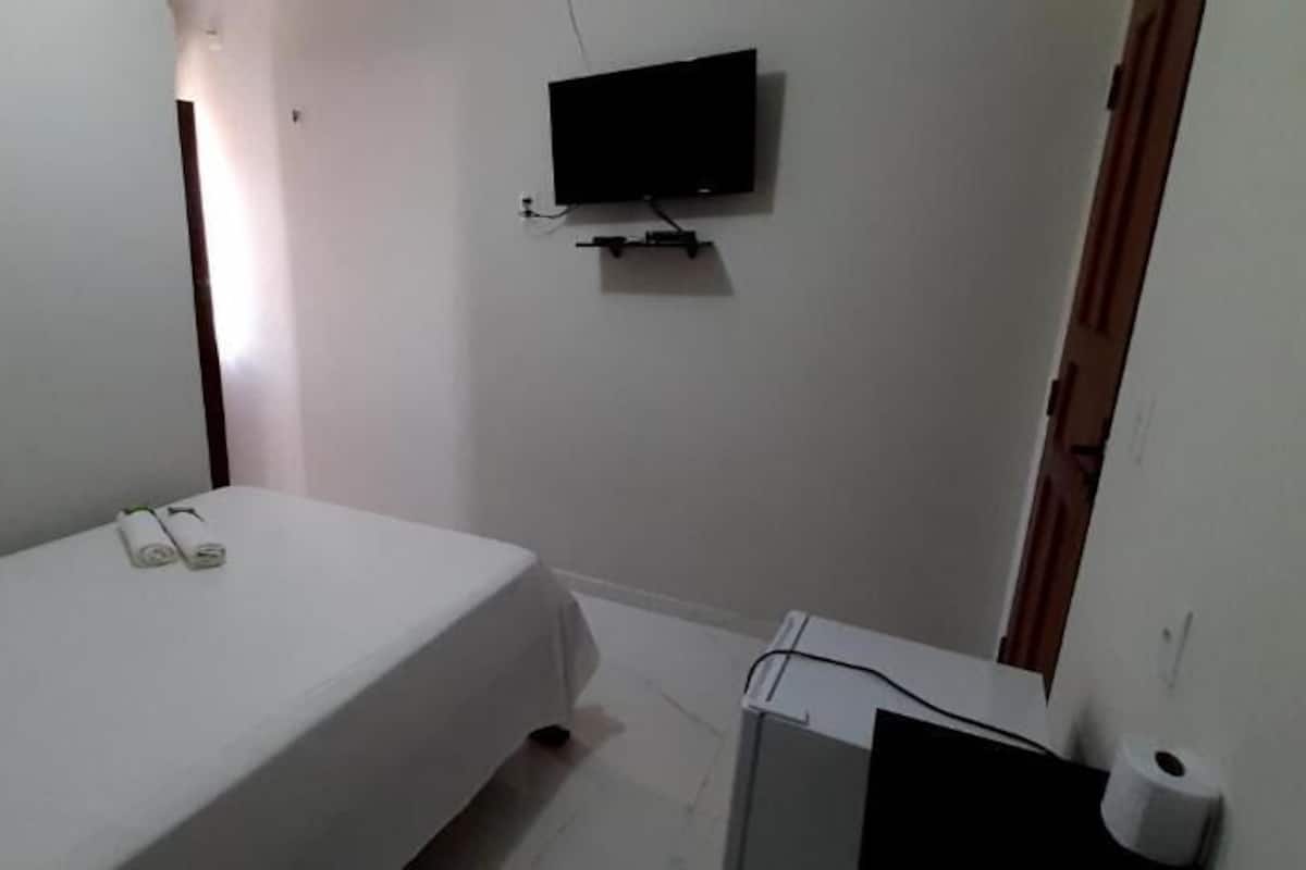 A small, sparsely furnished room with a single bed, a television mounted on the wall, and a door to the right.