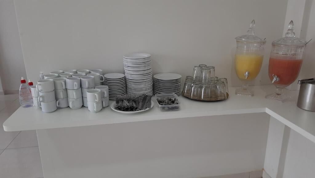 A buffet setup with stacks of white plates, bowls, and glasses, along with two dispensers of orange and red beverages.