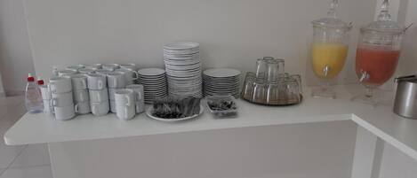A buffet setup with stacks of white plates, bowls, and glasses, along with two dispensers of orange and red beverages.
