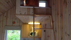 Une petite cabane en bois avec un espace de couchage en mezzanine, une kitchenette et une fenêtre donnant sur la verdure.
