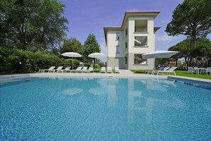 A swimming pool with lounge chairs and umbrellas, surrounded by trees and a multi-story building.