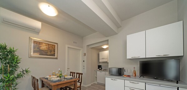 A modern kitchen with white cabinets, a dining table set for two, a television, and a plant. - Villa Caterina 2 by Wonderful Italy (Desenzano del Garda)