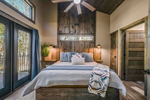 A bedroom with a large bed, wooden ceiling fan, two bedside lamps, a window with curtains, and a door.