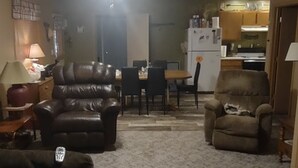 Una sala de estar con alfombra amarilla, dos sillones de cuero, una mesa de comedor con sillas, un ventilador de techo y una cocina al fondo.