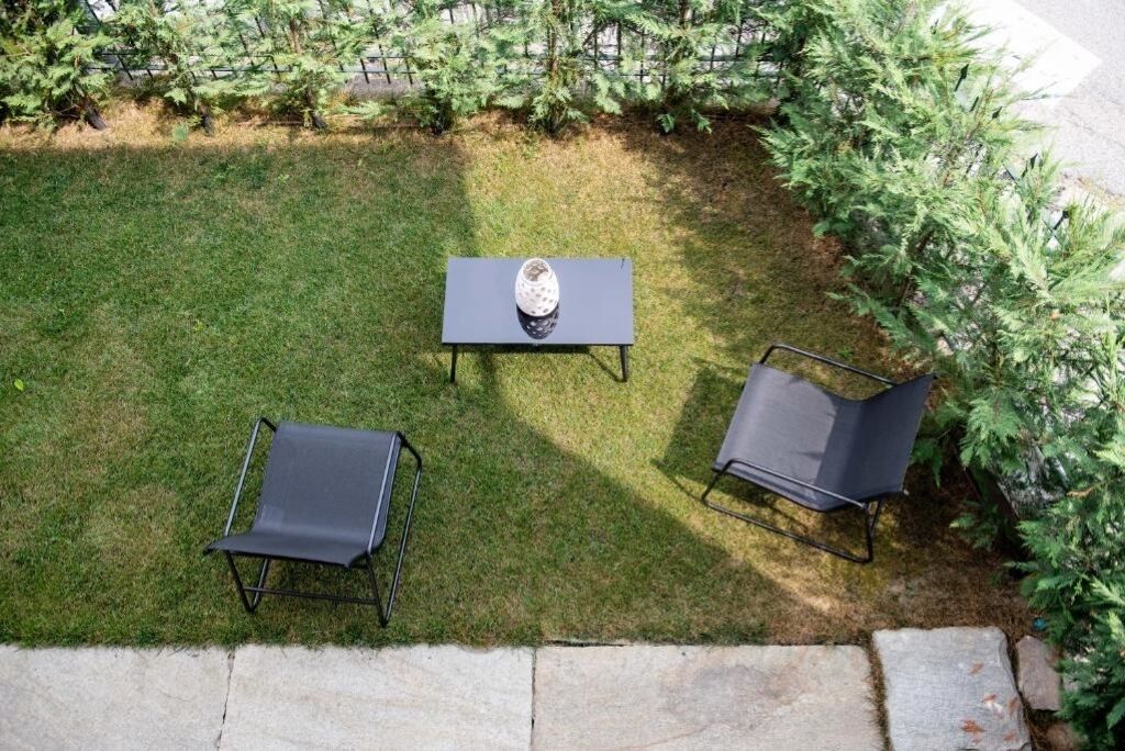 Uma área de pátio com uma mesa e duas cadeiras, rodeada por um gramado e folhagem densa.