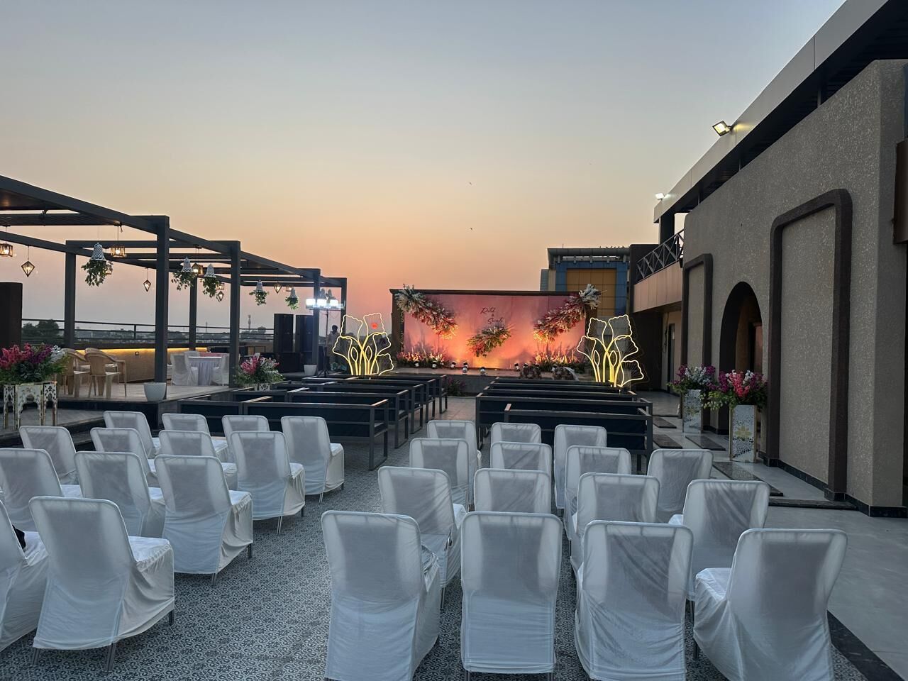 An outdoor seating area with rows of chairs, a stage with floral decorations, and a view of the sky.