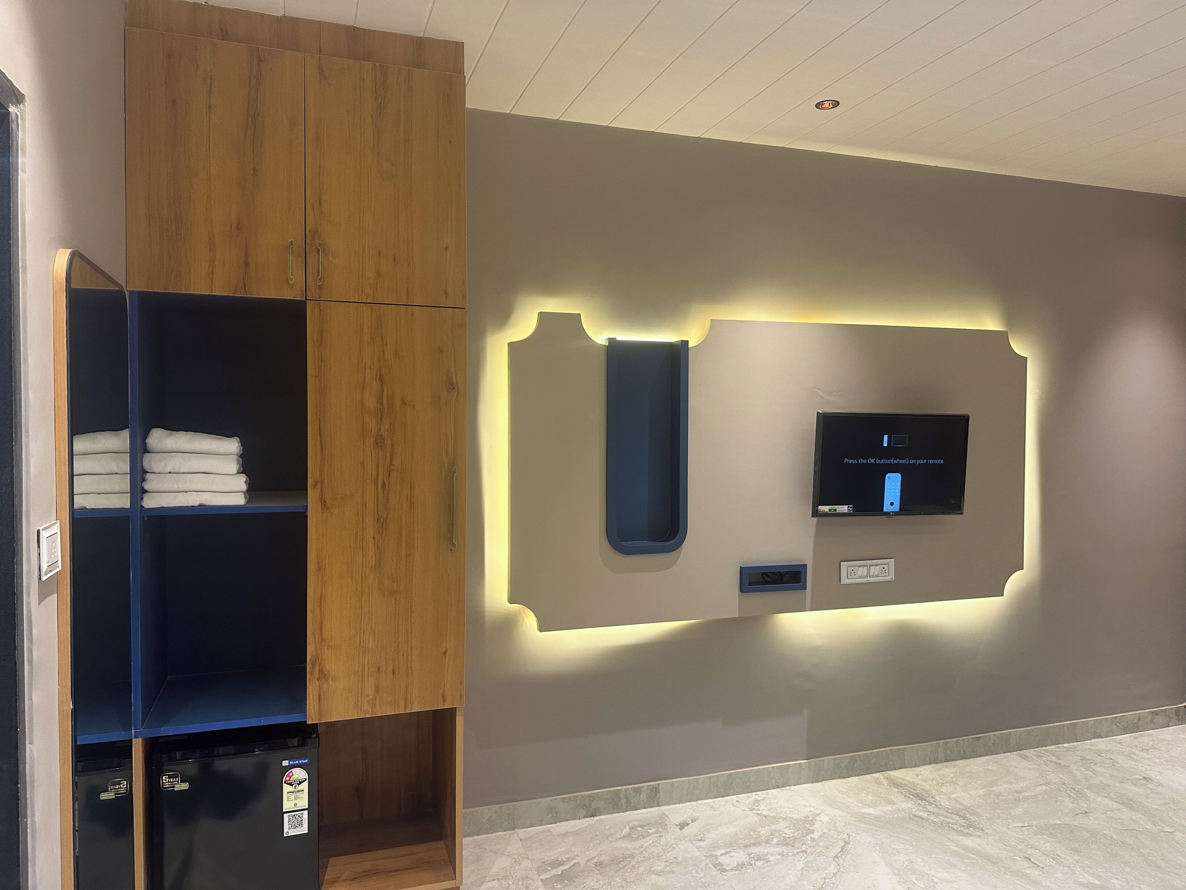 A hotel room with a wooden wardrobe, a wall-mounted TV, and a built-in shelf with folded towels.