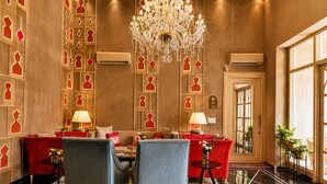 Een luxueuze kamer met een kroonluchter, rode en blauwe banken, een eettafel en een groot raam.
