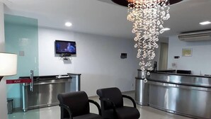 Un'area di accoglienza moderna con una TV a parete, una scrivania della reception, due sedie nere, un tavolino in vetro con un vaso di fiori e un lampadario.