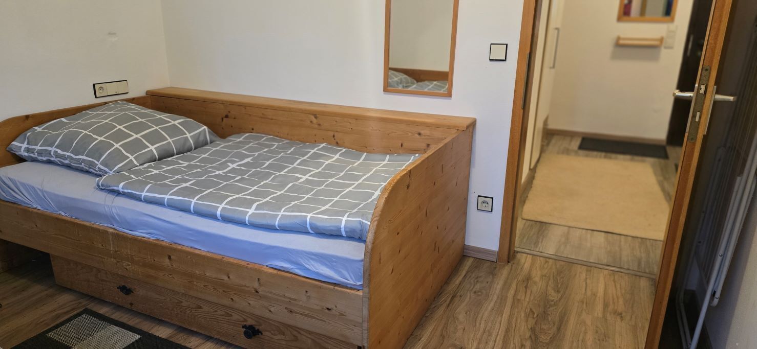 Una cama pequeña de madera con un colchón azul y dos almohadas, una mesita de noche de madera, un espejo y una puerta que da a otra habitación.