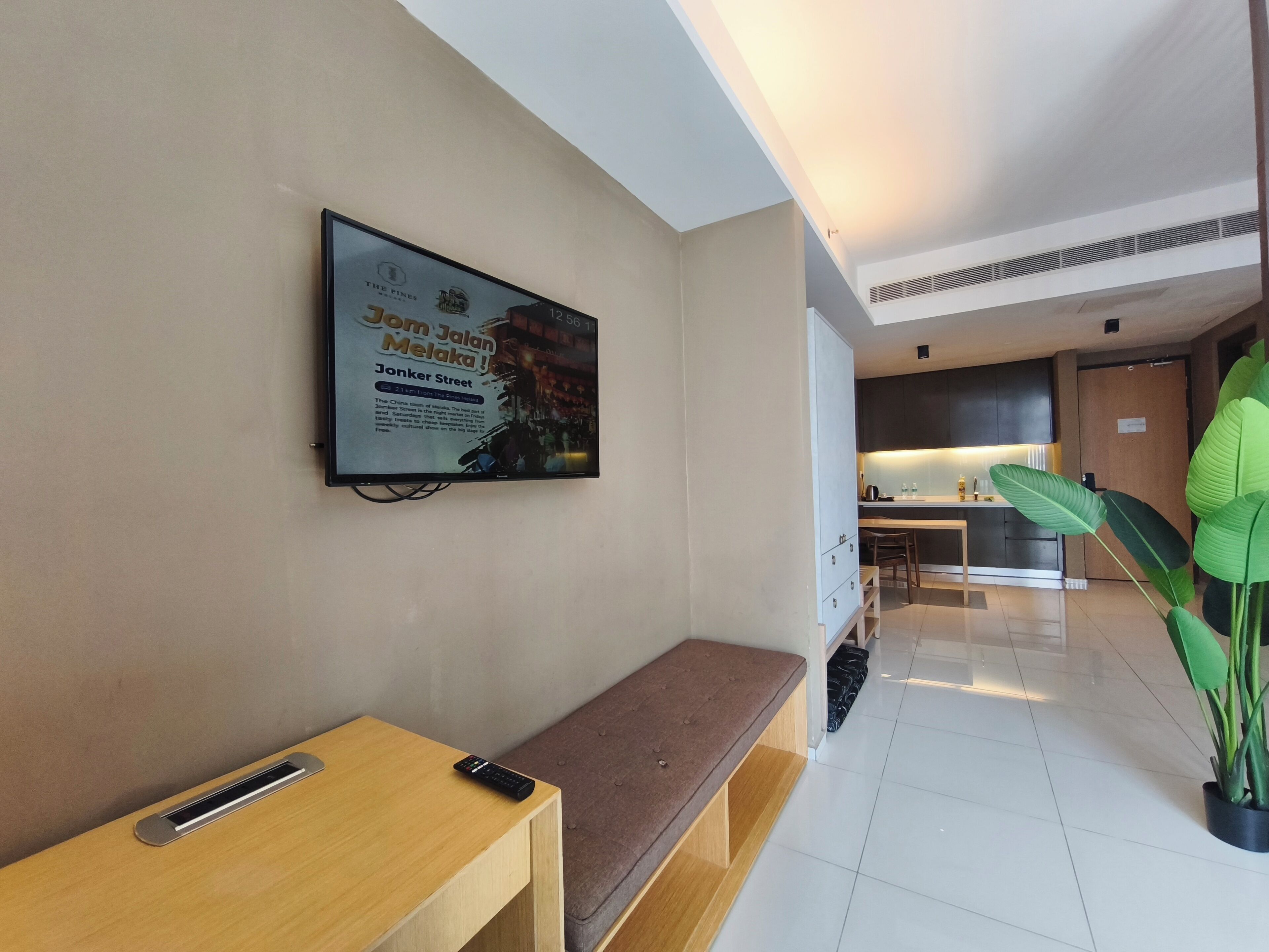 A modern hotel lobby with a TV mounted on the wall displaying an advertisement, a wooden bench, a remote control on the bench, a potted plant, and a glimpse of the kitchen area in the background.