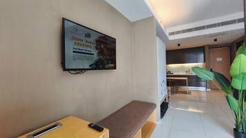 A modern hotel lobby with a TV mounted on the wall displaying an advertisement, a wooden bench, a remote control on the bench, a potted plant, and a glimpse of the kitchen area in the background.