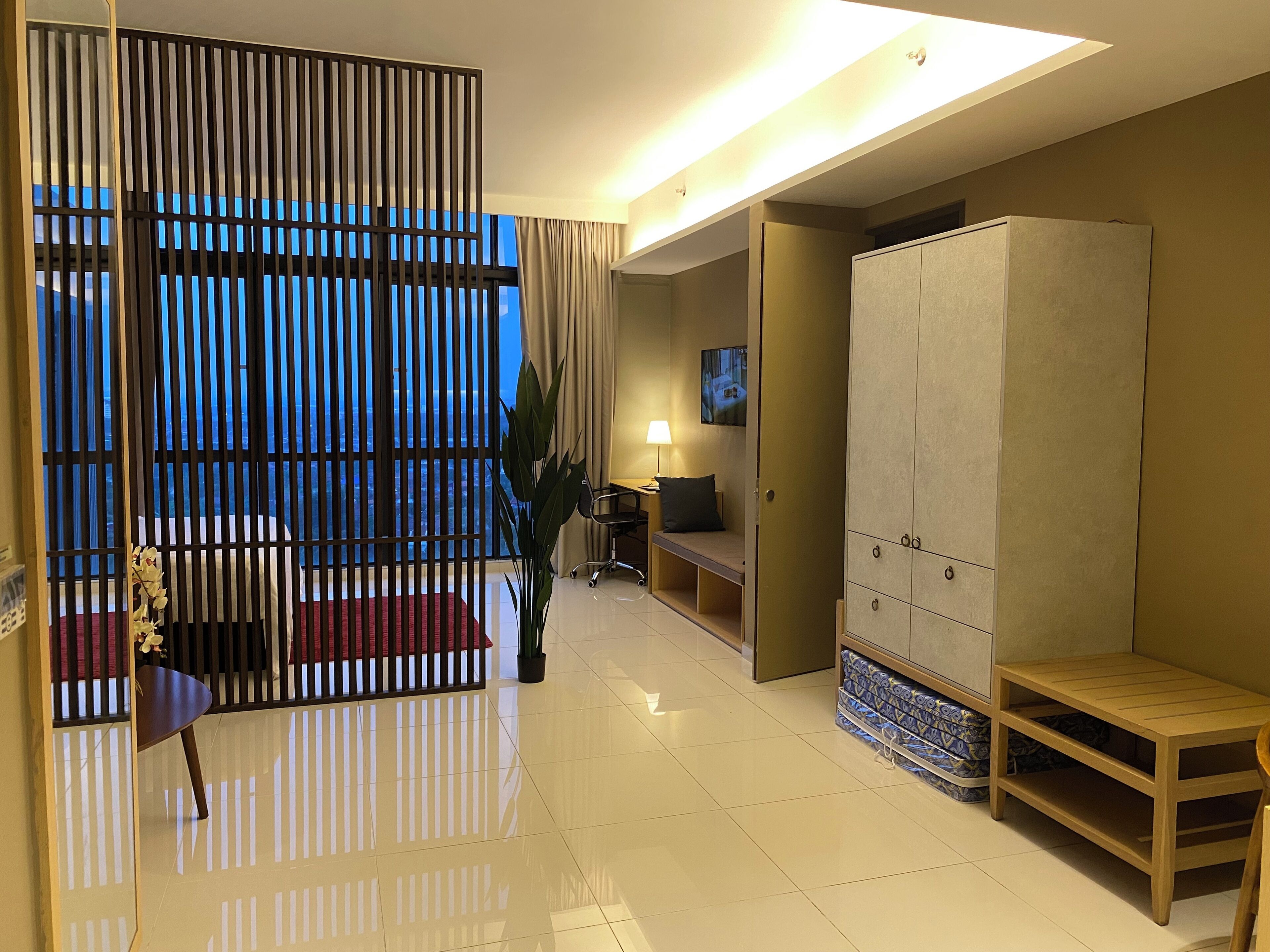 A modern hotel room with a sliding door leading to a balcony, a seating area with a bench, a wardrobe, and a television on the wall.