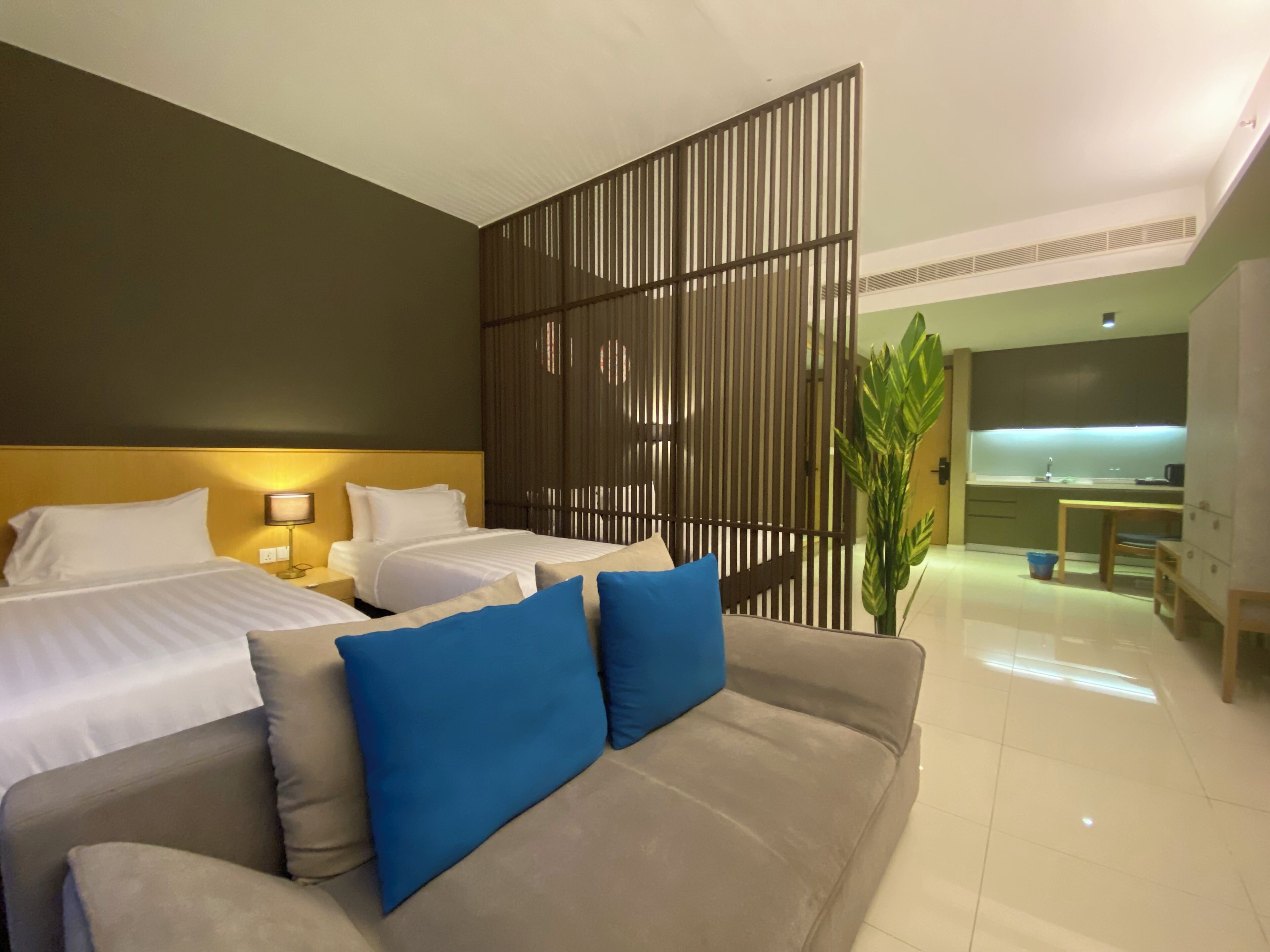 A hotel room with a large bed, a sofa with blue cushions, a partition wall, and a kitchenette in the background.