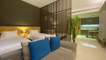 A hotel room with a large bed, a sofa with blue cushions, a partition wall, and a kitchenette in the background.