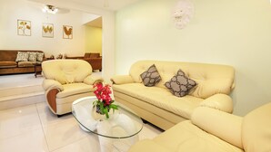 Una sala de estar moderna con un sofá beige, dos sillones, una mesa de centro de vidrio con flores y decoración en la pared.