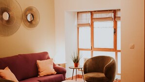 Un salon comprenant un canapé, un fauteuil et une petite table, un tapis à motifs et une fenêtre avec des stores.