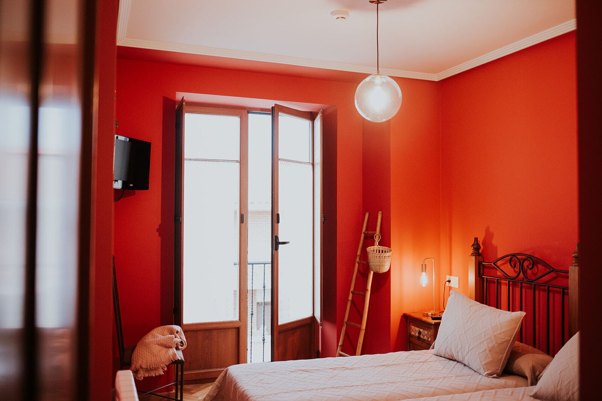 Une chambre avec un mur rouge, un lit avec des oreillers, une table de chevet, une lampe et une fenêtre donnant sur l’extérieur.