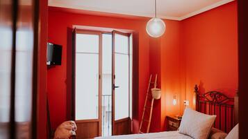 Une chambre avec un mur rouge, un lit avec des oreillers, une table de chevet, une lampe et une fenêtre donnant sur l’extérieur.