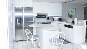 Una cocina moderna con armarios blancos, un refrigerador de acero inoxidable, un microondas y un lavamanos. Hay dos taburetes de bar blancos y un letrero que dice "Bon Appetit".