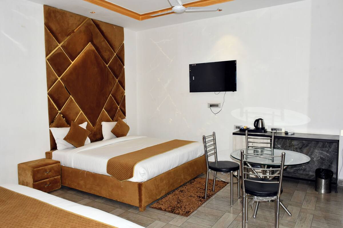 Habitación de hotel moderna con una cama grande, un televisor de pantalla plana en la pared, una zona de comedor con una mesa redonda y sillas, y una mesita auxiliar pequeña.