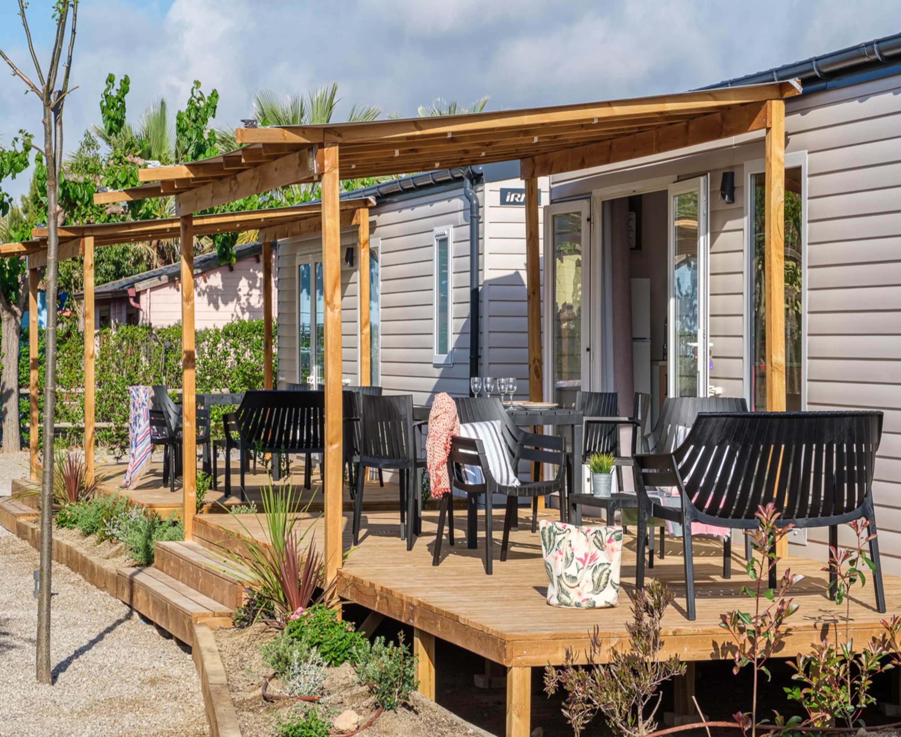 A covered outdoor seating area with wooden benches and tables, surrounded by plants and a gravel path.