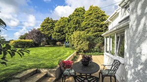 Une terrasse avec une table ronde et des chaises, un petit jardin avec une pelouse et des arbres, et un bâtiment blanc avec une fenêtre.