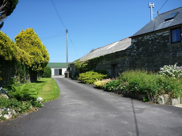 A narrow rural road flanked by stone buildings and lush greenery.