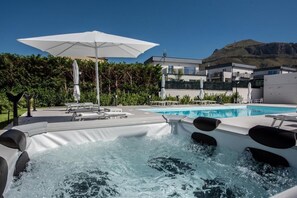Un espace avec un jacuzzi et une piscine, des parasols, et un bâtiment moderne en arrière-plan.